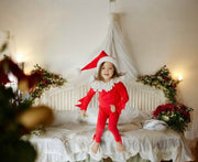 Child in a red elf costume sitting on a bed with Christmas decorations around.