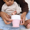 Child holding a pink cup with adult assistance, sitting on a white surface.