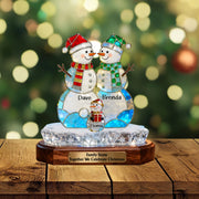 Decorative snowman family figure with personalized names on a wooden base, Christmas tree lights in the background.