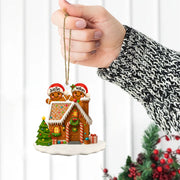 Hand holding a gingerbread house ornament with two gingerbread men wearing Santa hats against a white background.